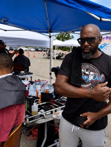 Volunteer barber standing beside grooming tools under a blue canopy